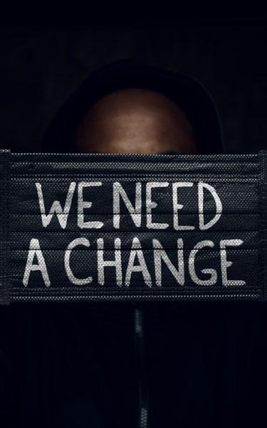 Black man holds up face mask with the words We need a change on it