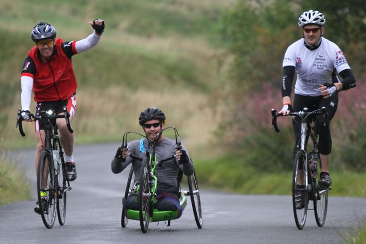 Cyclist with arm raised riding alongside a disabled rider on a customised 3 wheel bike