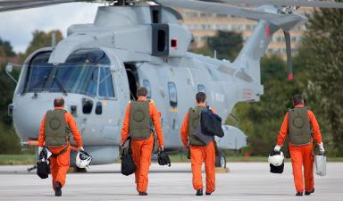 3 male pilots, dressed in orange flight suits and holding white helmets, walk towards an AW101 Merlin helicopter, with their backs turned to the camera