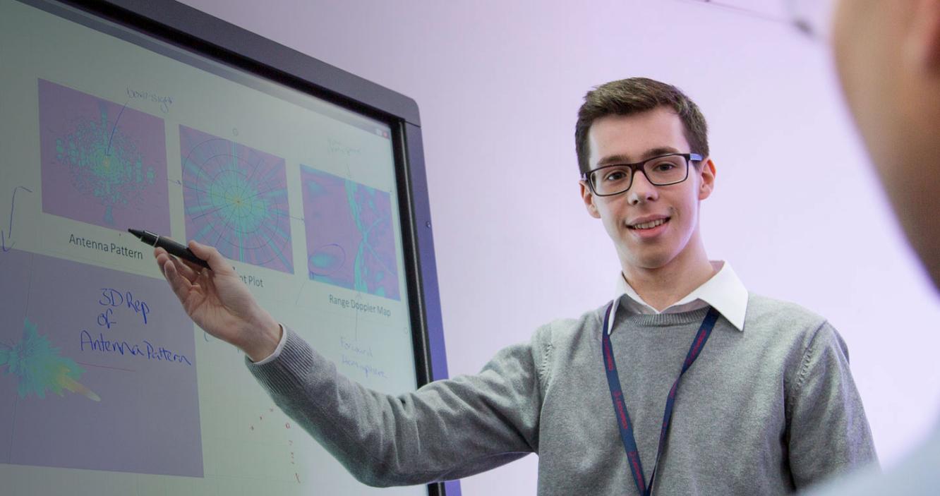 Young white male in glasses points to whiteboard and smiles