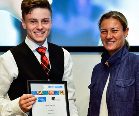 Young man in tartan tie and waistcoat smiles as he receives certificate from woman