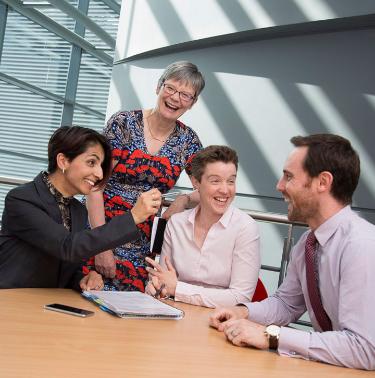 A group of colleagues, of mixed age, gender and ethnicity, laugh around a meeting room table