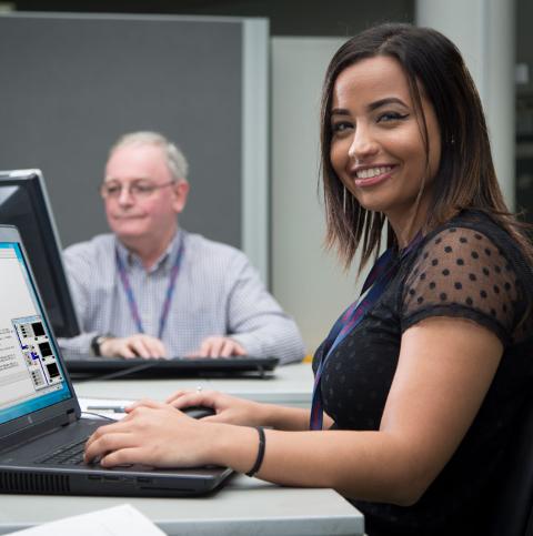Young woman sitting at desk with laptop turns and smiles to camera. Older man works on laptop in the background, out of focus.
