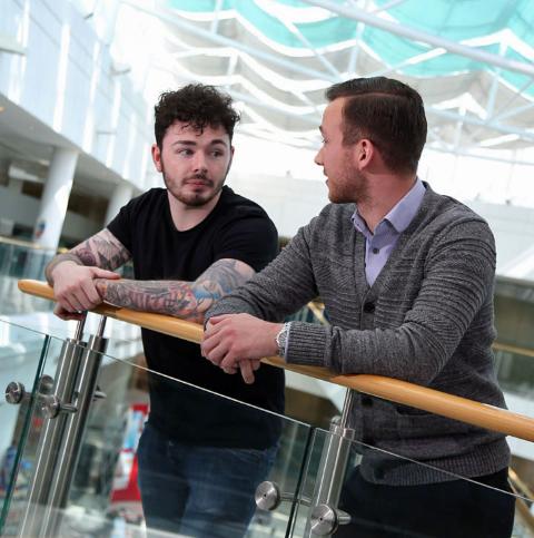 Two young men, one in a t shirt with tattoos, one in a shirt and cardigan, lean on balcony overlooking atrium at Leonardo site