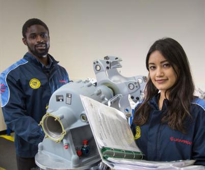 A young male and female engineer of different ethnicities smile and stand by machinery