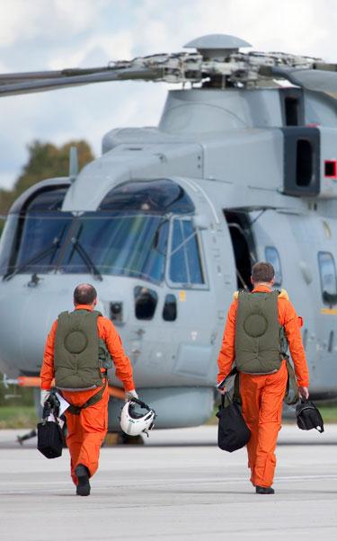 4 male pilots, dressed in orange flight suits and holding white helmets, walk towards an AW101 Merlin helicopter, with their backs turned to the camera
